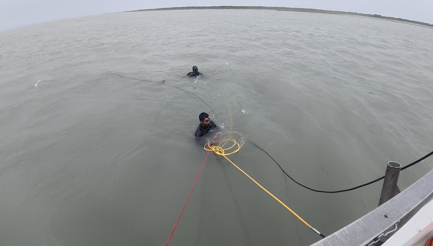 Two divers work in open gray water beside a boat with red, yellow, and black lines trailing between them.