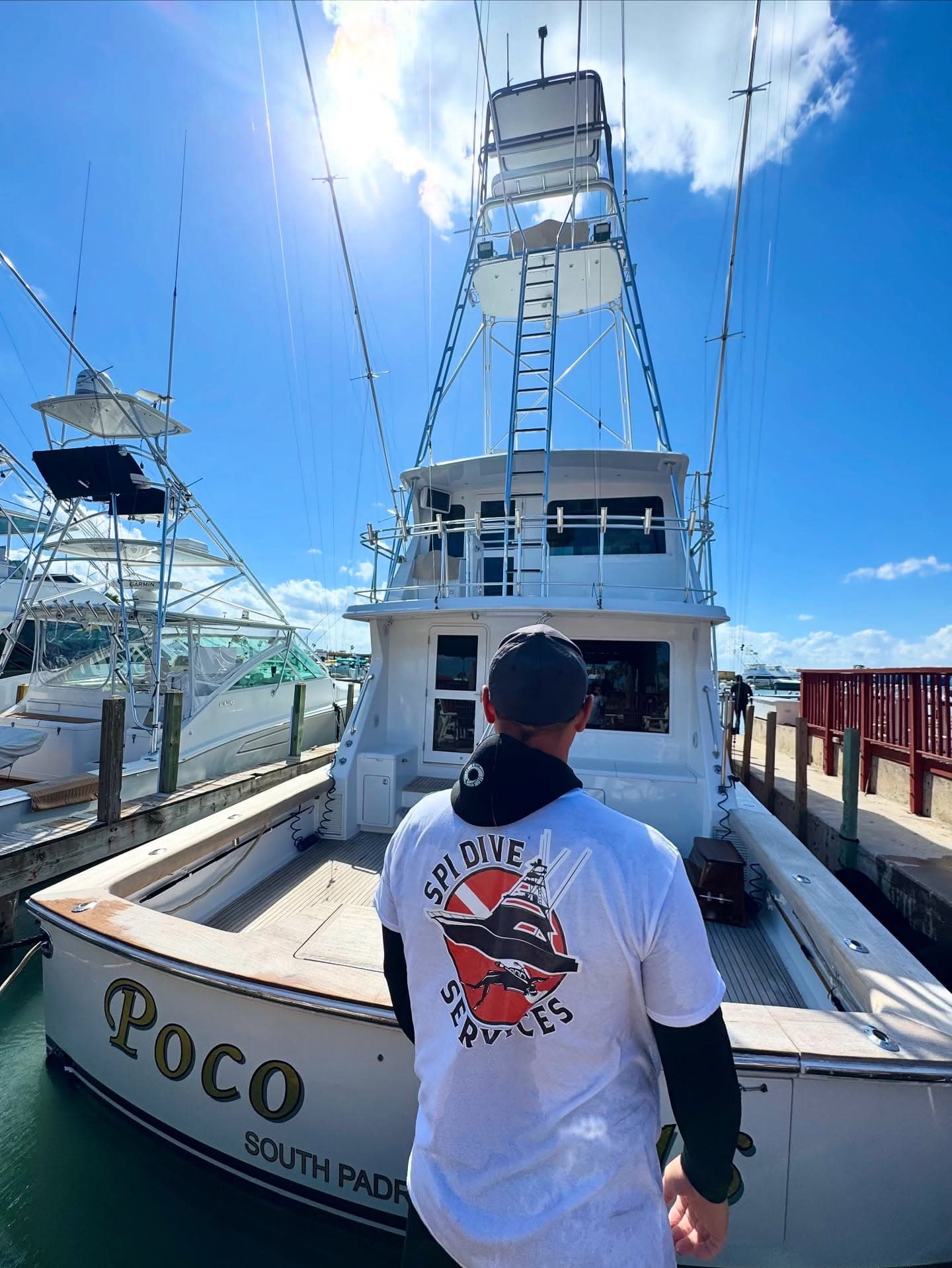 SPI Dive Services crew member approaches the stern of the sportfishing boat Poco Mas under a bright blue sky.